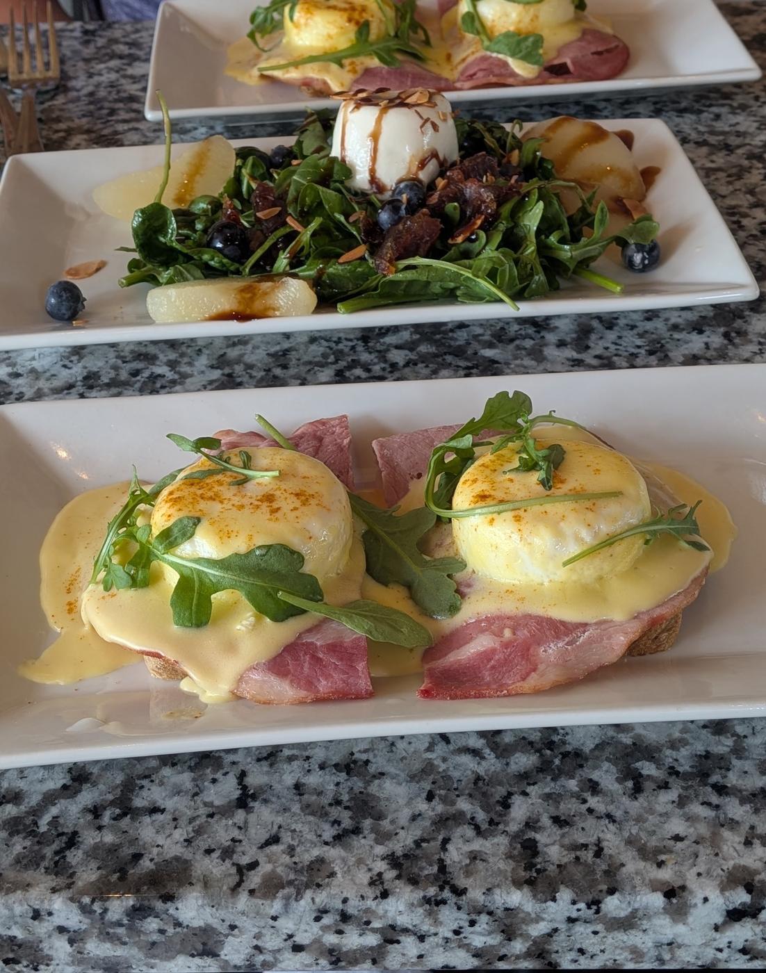 Two plates of Eggs Benedict with hollandaise, arugula and thick cut ham, either side of a fig and pear salad with blueberries, pear slices, burrata and toasted almonds.