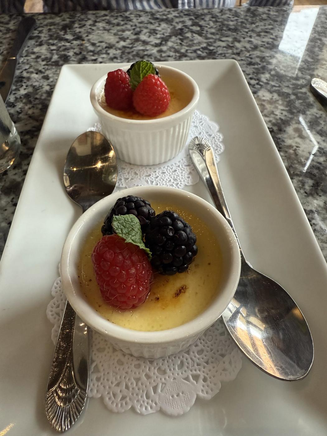 Two ramekins of a torched custard dessert topped with raspberries, blackberries and mint, on a white plate with lace doilies and silver spoons.