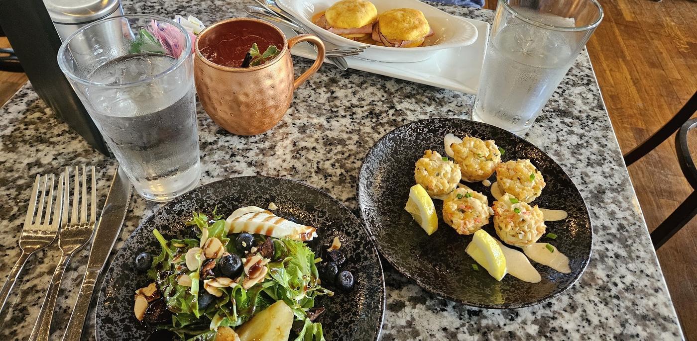 A laid table with the fig and pear salad, crab cakes with lemon, Eggs Benedict on biscuits, a copper mug and water glasses on a speckled stone table.